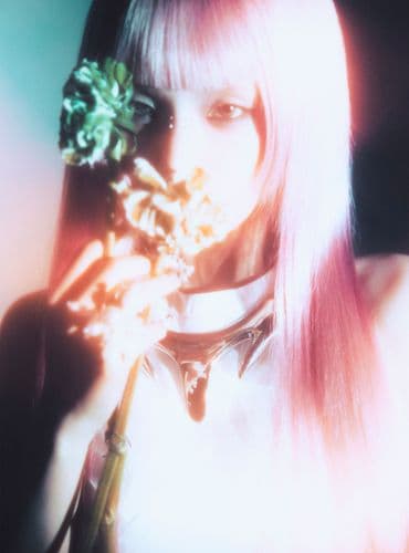 Woman with long pink hair holds a bouquet of small flowers near her face, soft lighting.
