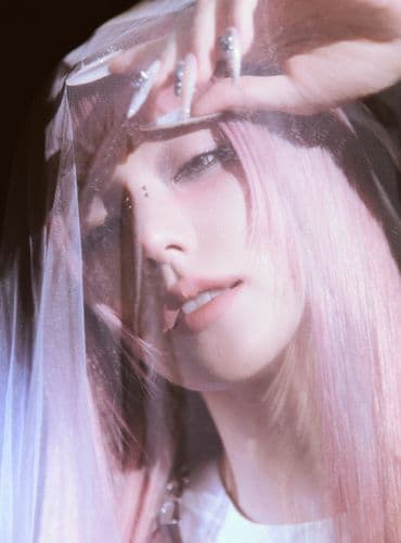 Close-up of a woman with pink hair, hand near her forehead, looking intently.