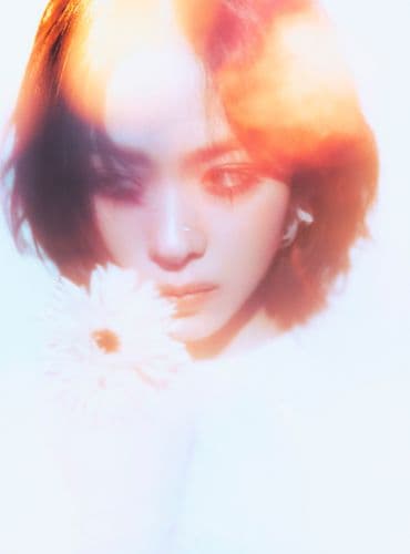 Close-up of a woman's face holding a white flower, soft colorful hazy lighting.